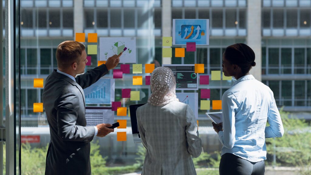 Three business professionals discussing charts and notes on a glass wall covered with colorful sticky notes.