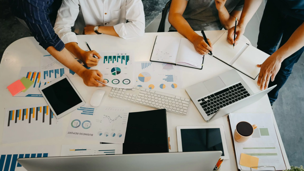 Overhead view of a business meeting with four people analyzing charts and using digital devices at a white table.