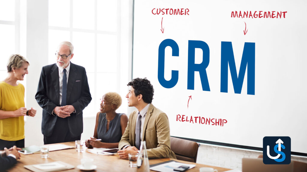 Four people in business attire are having a discussion in a conference room. A presentation slide behind them displays "CRM" with arrows labeled "Customer," "Management," and "Relationship.