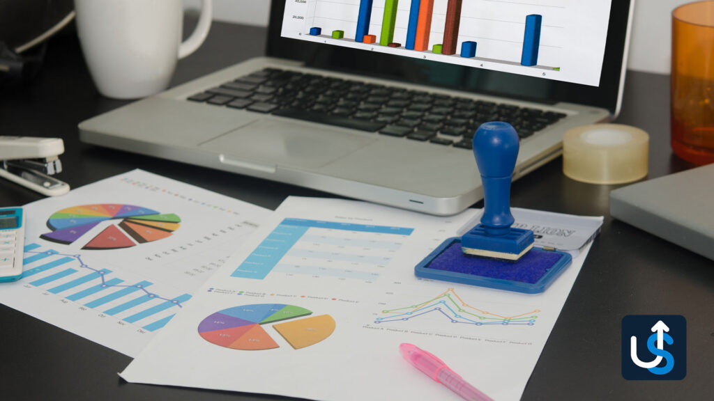 A laptop displaying graphs and charts is on a desk alongside printed charts, a blue ink stamp, a pink pen, a stapler, and an orange cup. Office supplies are scattered around.