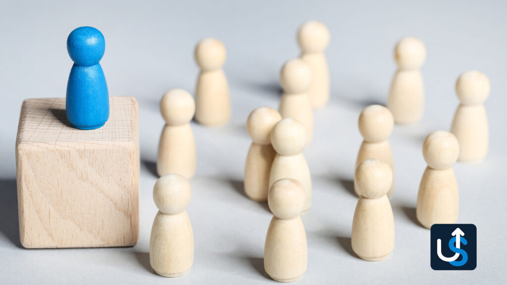 A blue wooden figure stands on a block above a group of beige wooden figures. A logo with an upward arrow is in the bottom-right corner.
