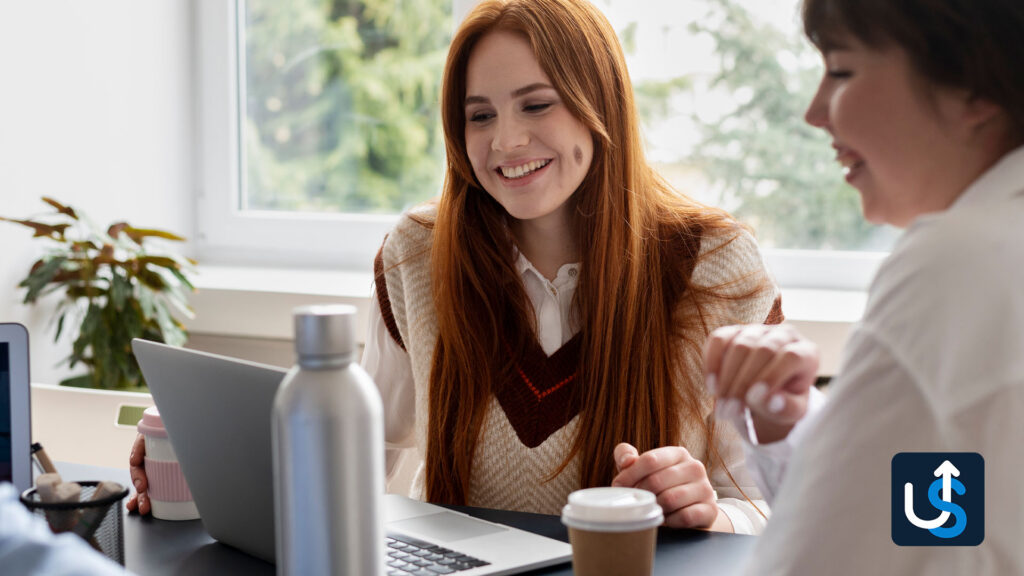 Two women sitting at a table, one with a laptop, smiling and talking. A plant and a coffee cup are on the table, and a window with green foliage is in the background.