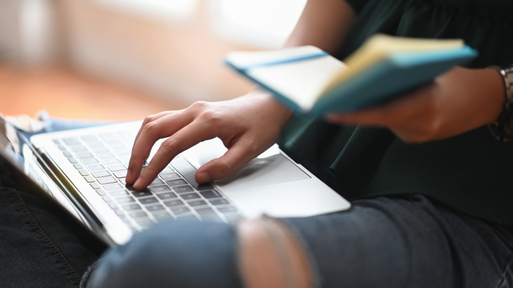 Person typing on a laptop with one hand while holding an open book in the other hand.