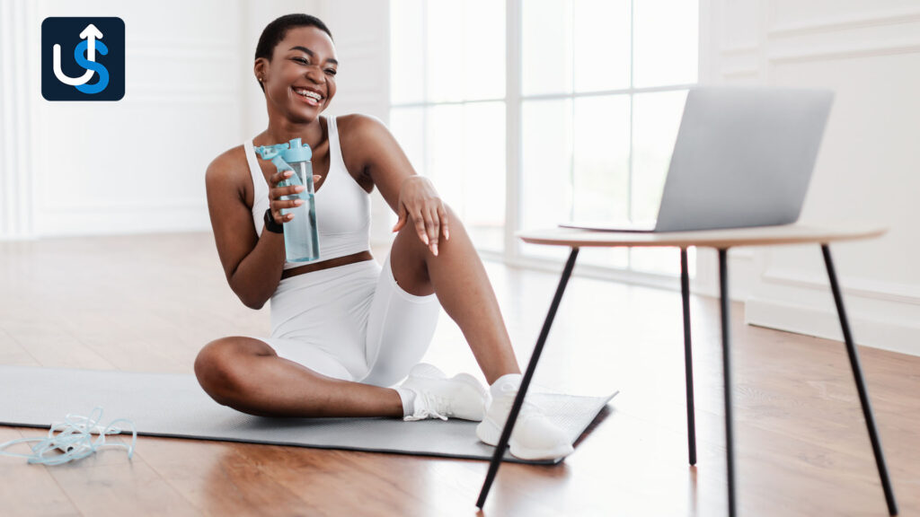 A person in workout attire sits on a mat, holding a water bottle, smiling at a laptop on a small table in a bright room.
