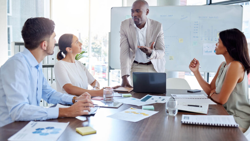 Four people in a meeting room having a discussion, with a man standing near a whiteboard covered with charts and notes.