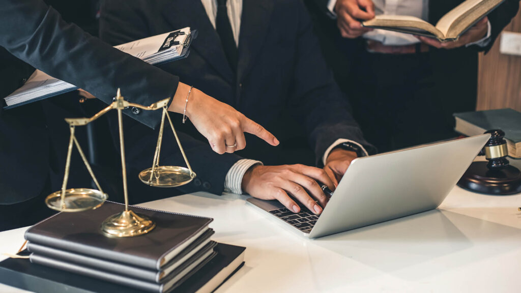 A person points at a laptop screen as another types. A scale, books, and a gavel are on the table, suggesting a legal setting.