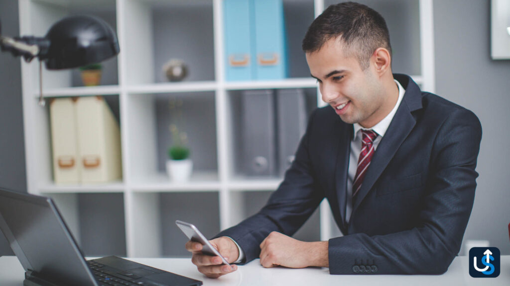 A man in a suit smiles while looking at his phone, seated at a desk with a laptop and shelves in the background.