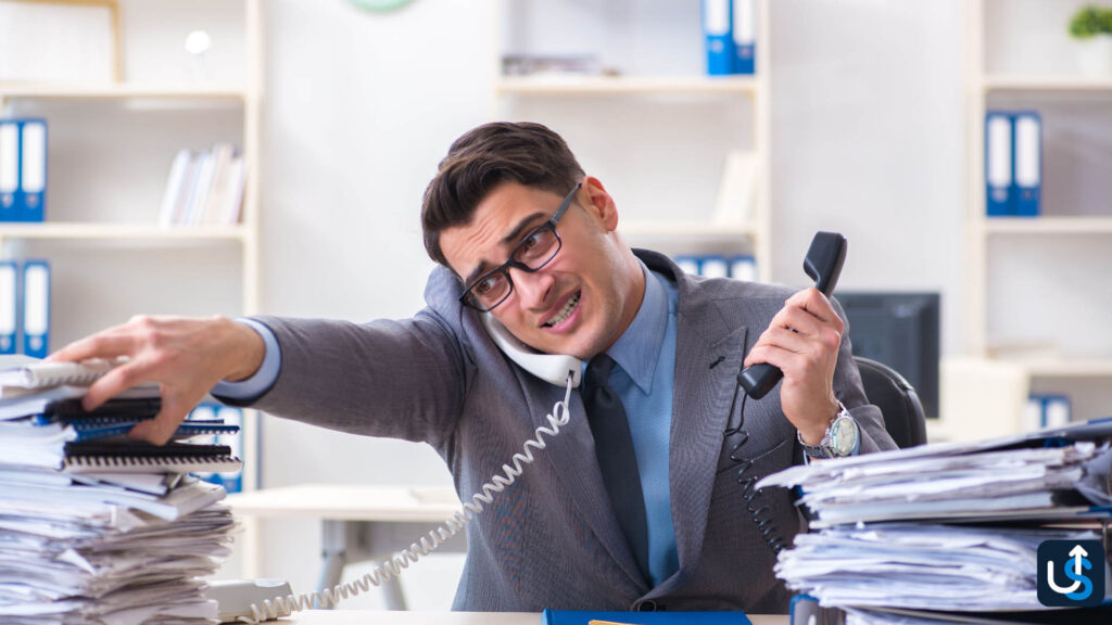 A man in an office suit multitasks, using a phone while reaching for a stack of papers on his cluttered desk.