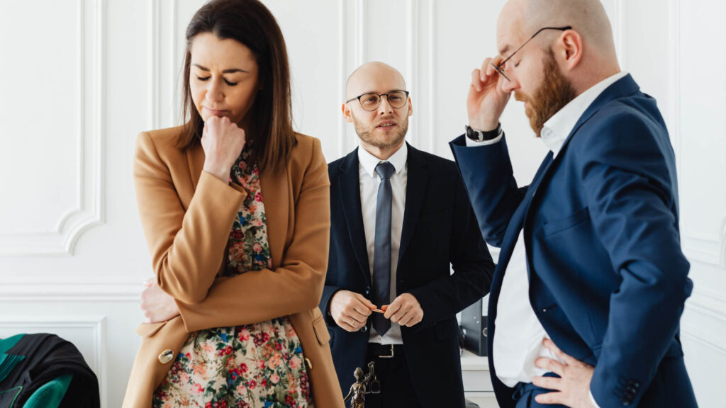 Three people in business attire are engaged in a thoughtful discussion in a bright office setting.