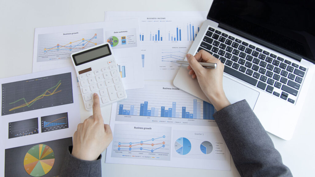 Person analyzing printed graphs and charts with a calculator and a laptop on a desk, reviewing business income and growth data.