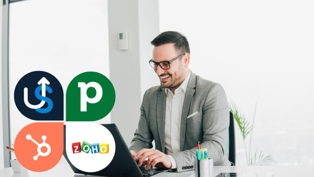 A man in a suit works on a laptop at a desk with CRM software logos including HubSpot, Zoho, Pipedrive, and UpLead displayed in the foreground.